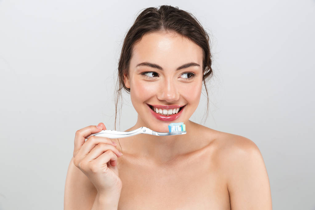 Retrato de beleza de uma jovem mulher de topless sorridente com maquiagem segurando escova de dentes com pasta de dentes isolada sobre fundo cinza
 - Foto, Imagem