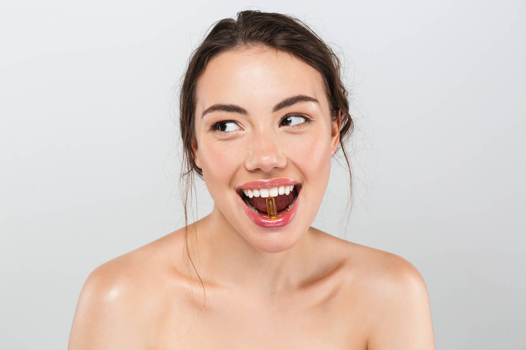 Retrato de belleza de una joven alegre en topless con maquillaje sosteniendo una píldora en su boca aislada sobre fondo gris
 - Foto, Imagen