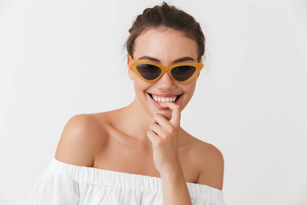 Πορτρέτο του μια χαρούμενη περιστασιακή μελαχρινή νεαρή γυναίκα σε γυαλιά ηλίου που βλέπουν τα φωτογραφικών μηχανών που απομονώνονται σε λευκό φόντο - Φωτογραφία, εικόνα