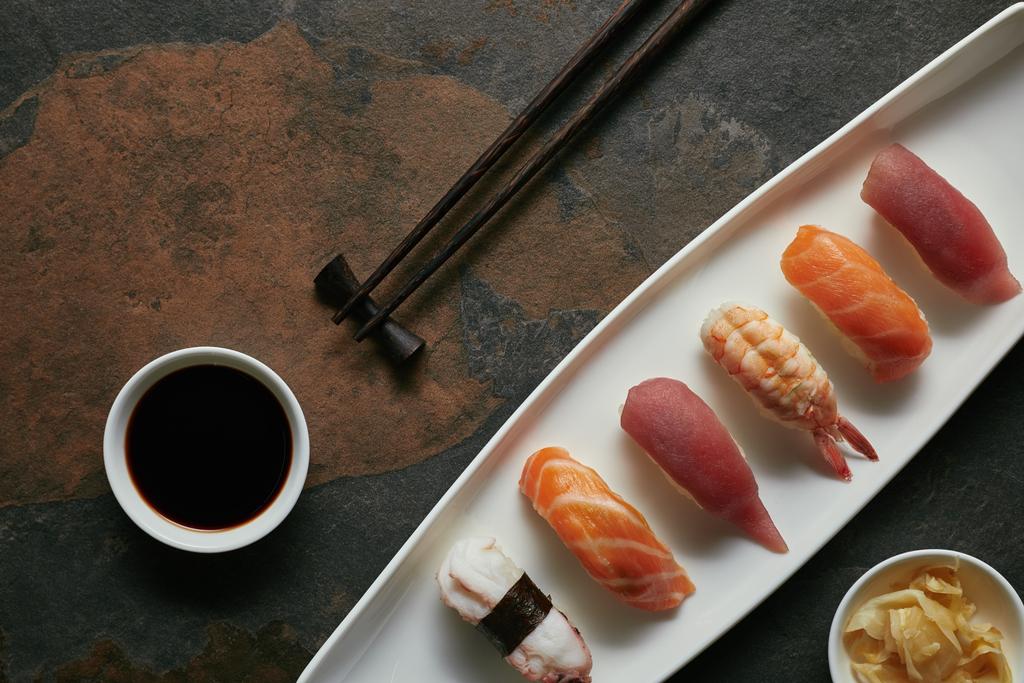 top view of assorted nigiri sushi set, chopsticks and soya sauce on white plate on dark surface - Photo, Image