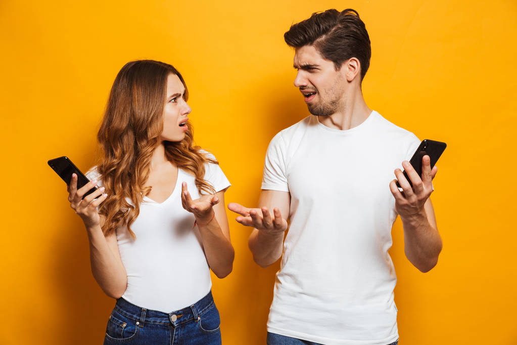 Photo of discontent man and woman looking at each other with perplexity while holding mobile phones isolated over yellow background - Photo, Image