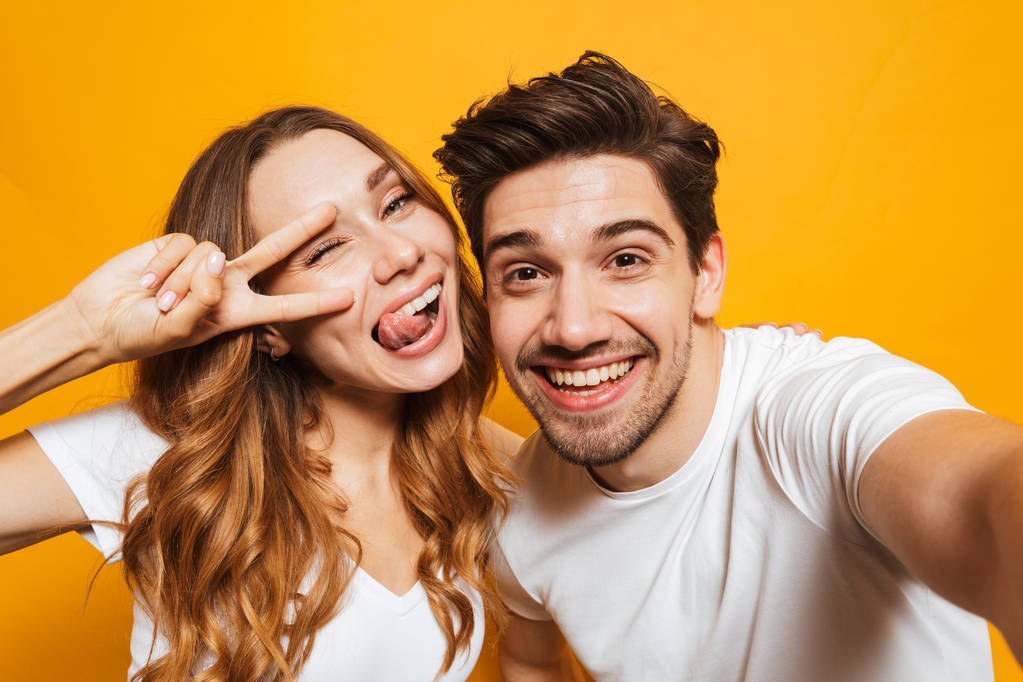 Porträt eines lächelnden jungen Paares, das ein Selfie macht und ein Friedenszeichen isoliert auf gelbem Hintergrund zeigt - Foto, Bild