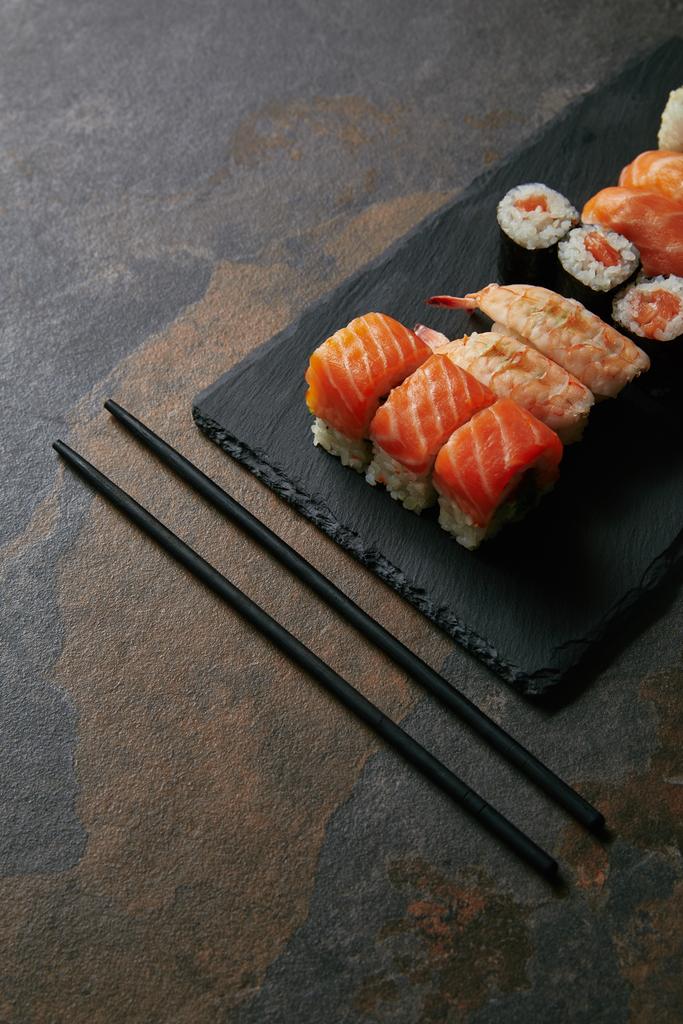 κλείνω πάνω θέα τοποθετημένα σούσι σε μαύρη πλάκα πλάκα και chopsticks σε σκούρα επιφάνεια - Φωτογραφία, εικόνα
