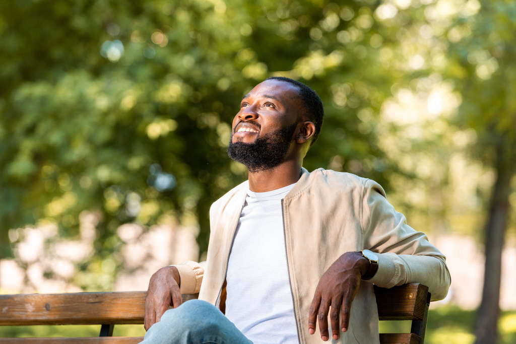 przystojny uśmiechający się african american człowiek siedzi na drewnianej ławce w parku i patrząc w górę  - Zdjęcie, obraz