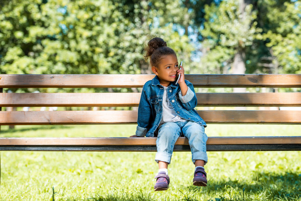 rozkošný afroamerické kid sedí na lavičce v parku a mluví o smartphone - Fotografie, Obrázek