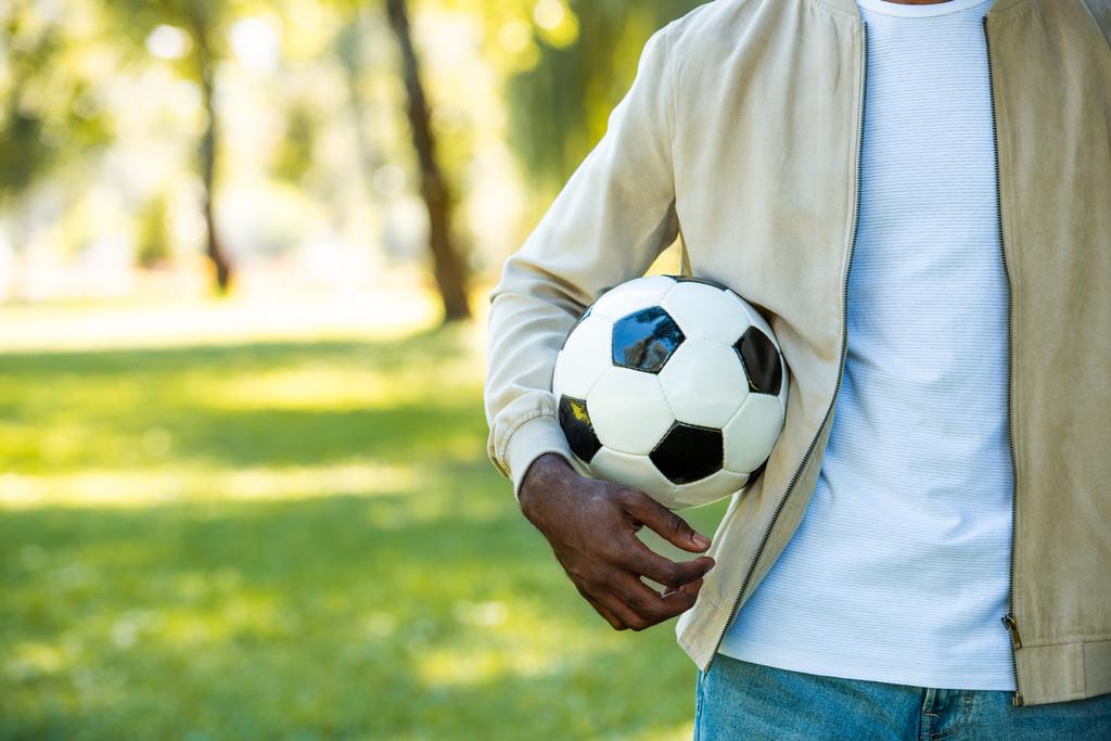 bijgesneden afbeelding van Afro-Amerikaanse man die houdt van voetbal bal in park - Foto, afbeelding