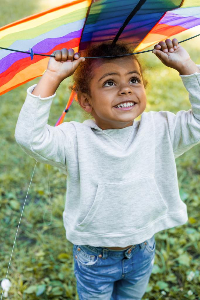 smiling adorable african american kid holding rainbow kite above head in park - Photo, Image