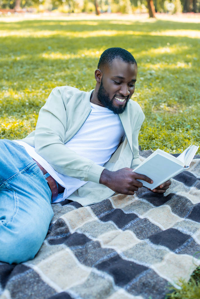 χαμογελώντας όμορφος αφρικανική αμερικανική άνθρωπος ξαπλωμένος σε κουβέρτα στο πάρκο και την ανάγνωση βιβλίων - Φωτογραφία, εικόνα