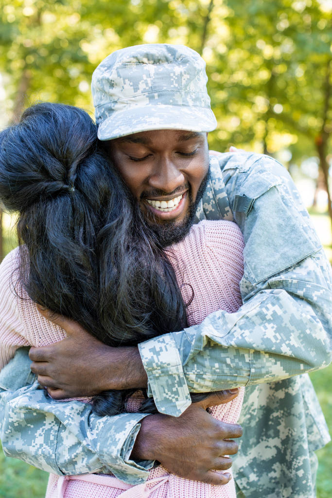 glimlachend Afrikaanse Amerikaanse soldaat in militaire uniform knuffelen vriendin in park - Foto, afbeelding