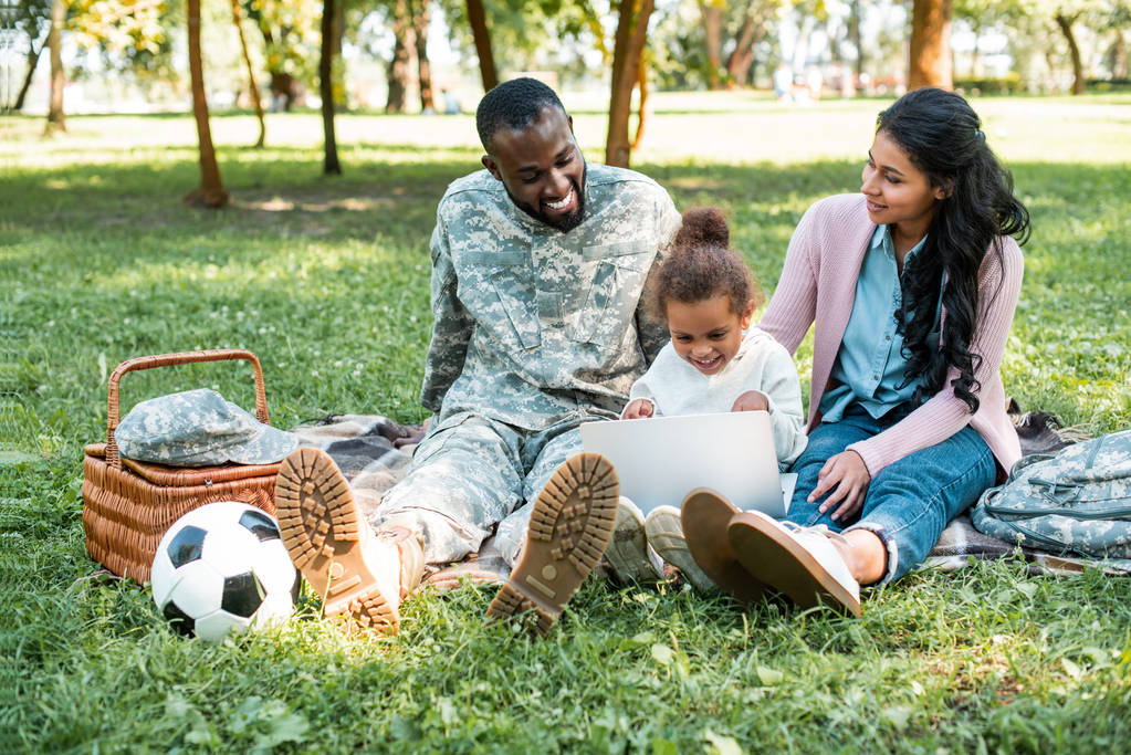 soldado afroamericano en uniforme militar mirando cómo hija usando portátil en parque
 - Foto, Imagen