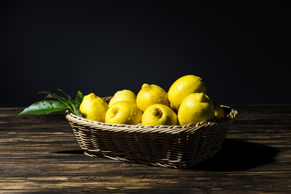 gelbe Zitronen mit Blättern im Weidenkorb auf Holztisch mit schwarzem Hintergrund - Foto, Bild