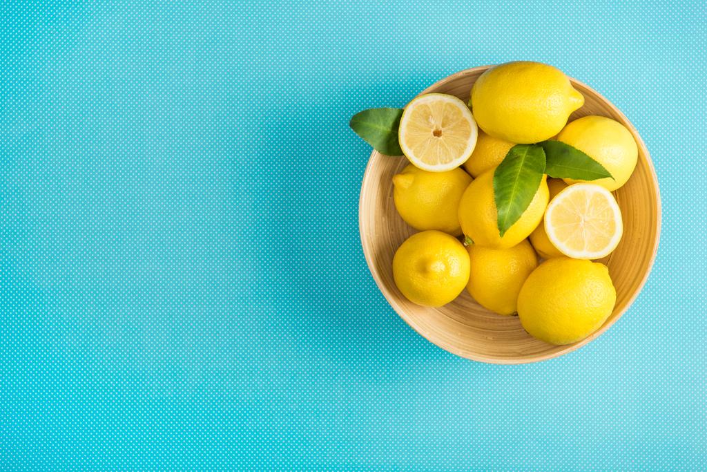 Draufsicht auf frische Zitronen mit Blättern in Holzteller auf türkisfarbenem Hintergrund - Foto, Bild