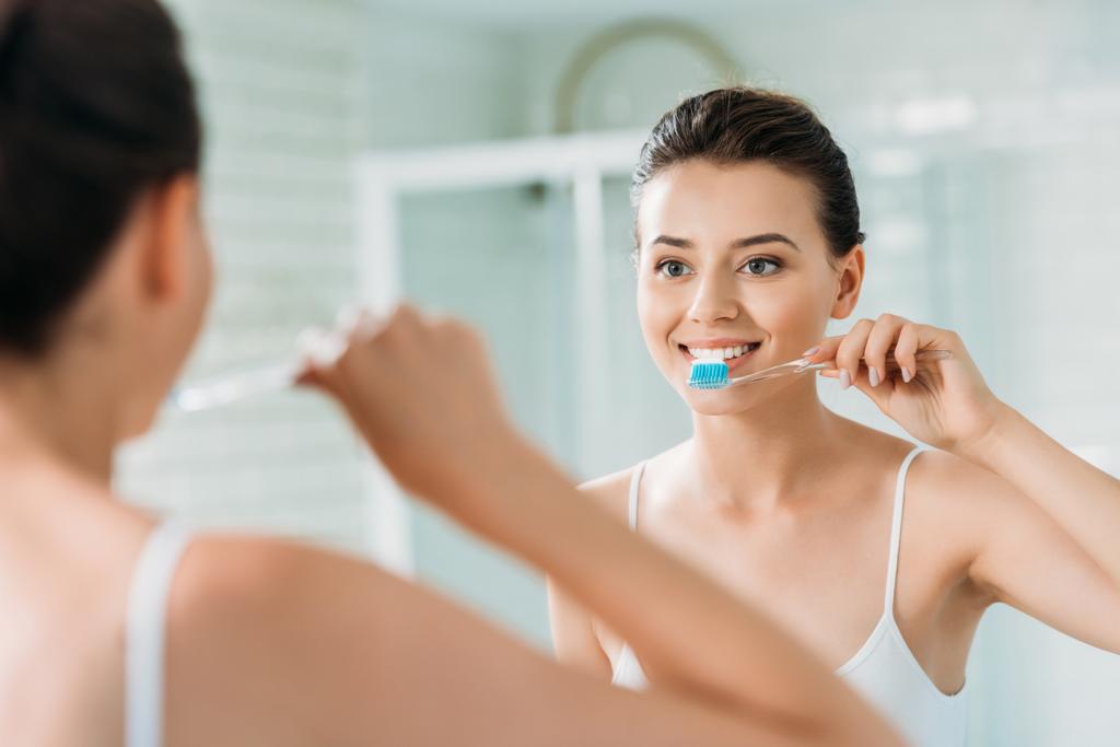 красивая улыбающаяся девушка чистит зубы перед зеркалом в ванной комнате
 - Фото, изображение