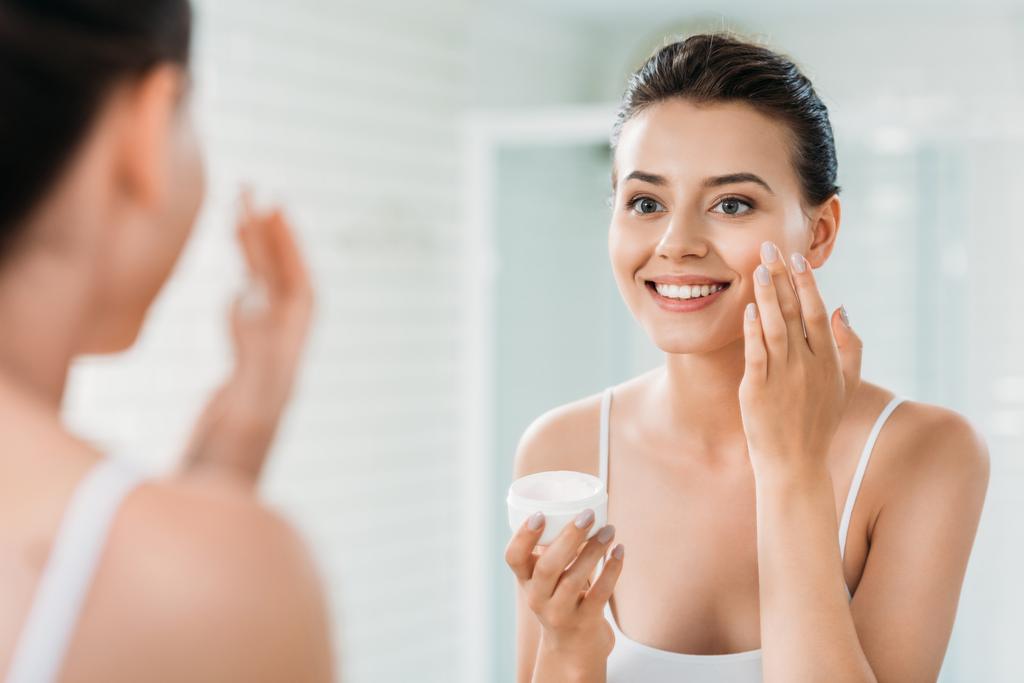 mooi blij meisje toe te passen gezicht crème en op zoek naar spiegel in de badkamer  - Foto, afbeelding