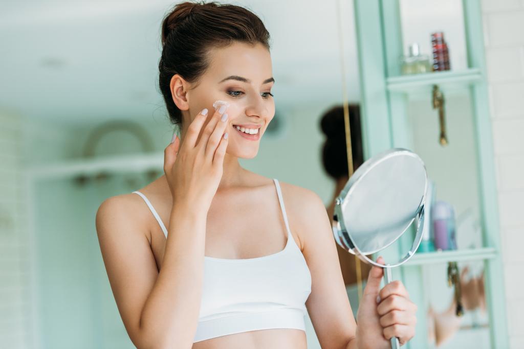 красива усміхнена дівчина тримає дзеркало і наносить крем для обличчя у ванній
  - Фото, зображення