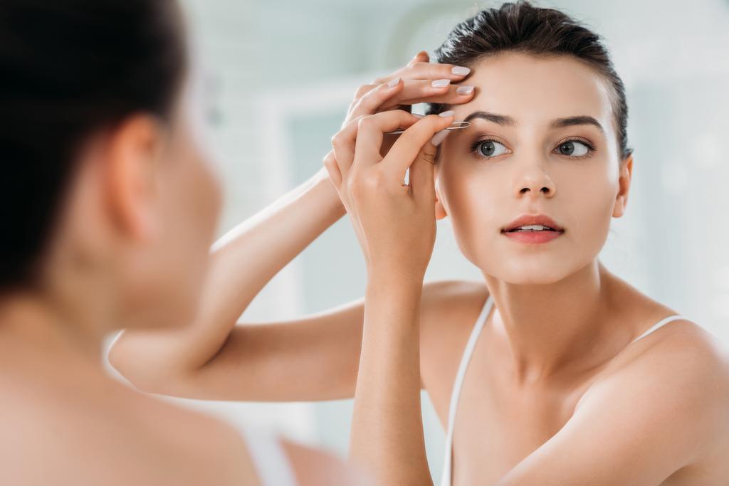 schöne Mädchen korrigiert Augenbrauen mit Pinzette und Blick auf Spiegel im Badezimmer  - Foto, Bild