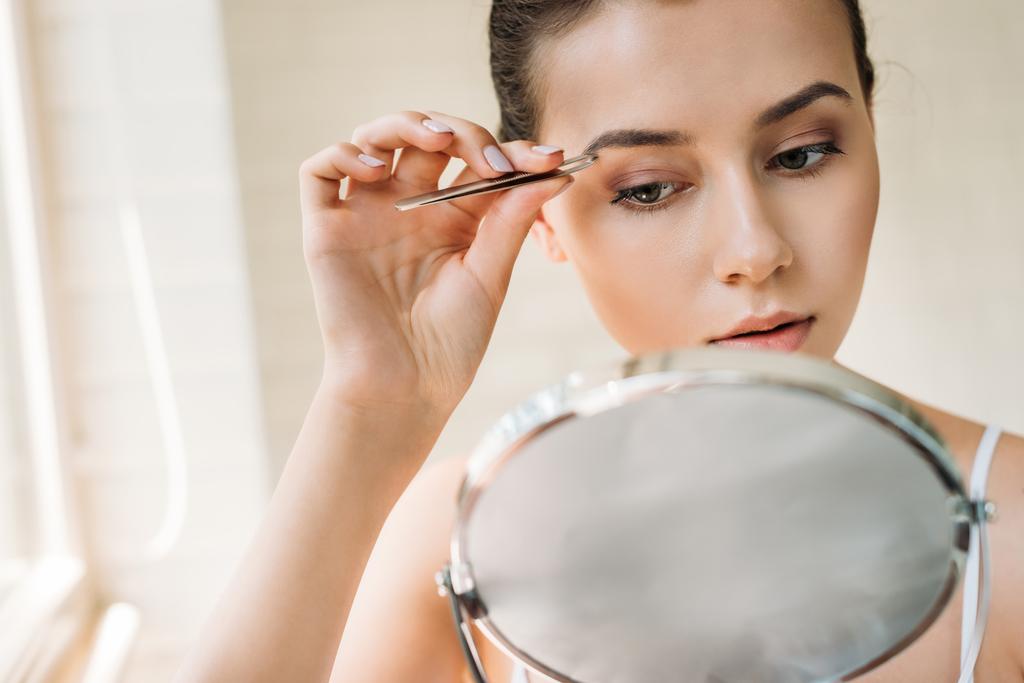 mulher atraente corrigindo sobrancelhas com pinças e olhando para o espelho
   - Foto, Imagem