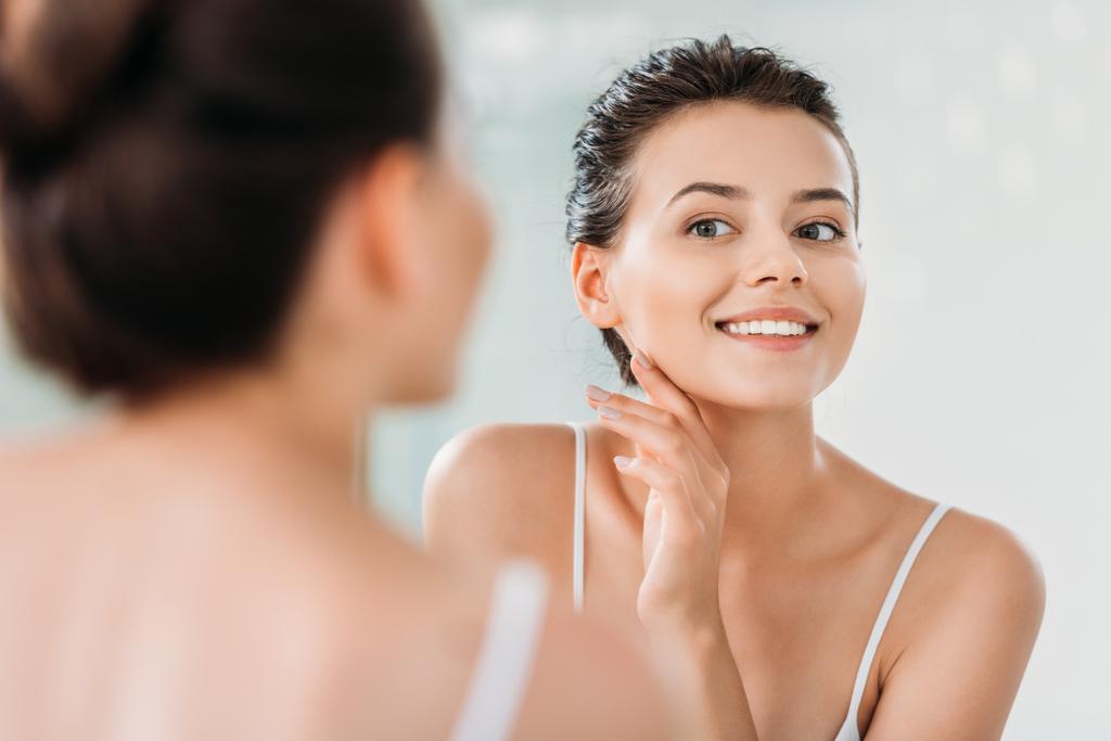 piękna, uśmiechnięta młoda kobieta dotyka skóry i patrząc w lustro w łazience - Zdjęcie, obraz