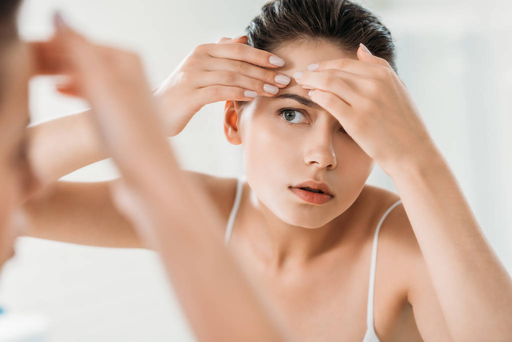 επιλεκτική εστίαση νεαρής γυναίκας τον έλεγχο του δέρματος στο μέτωπο και κοιτάζοντας καθρέφτη στο μπάνιο   - Φωτογραφία, εικόνα