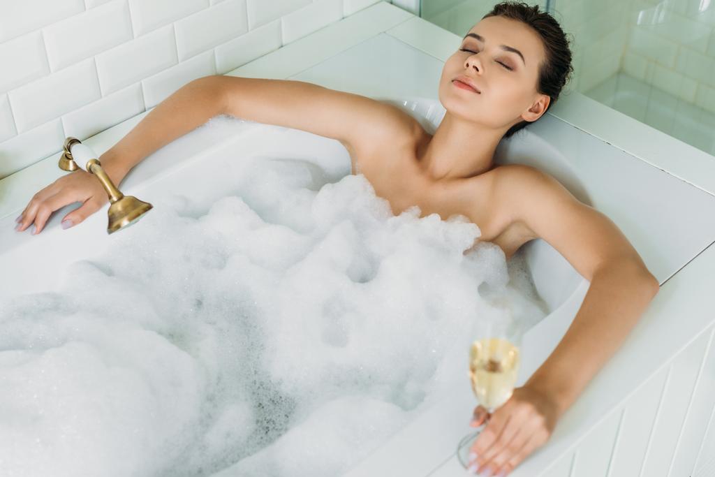 Hochwinkelaufnahme der schönen jungen Frau mit geschlossenen Augen, die ein Glas Champagner hält und sich in der Badewanne mit Schaum entspannt - Foto, Bild