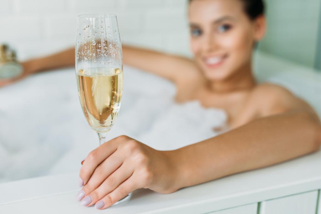 close-up view of beautiful smiling young woman holding glass of champagne in bathtub - Photo, Image