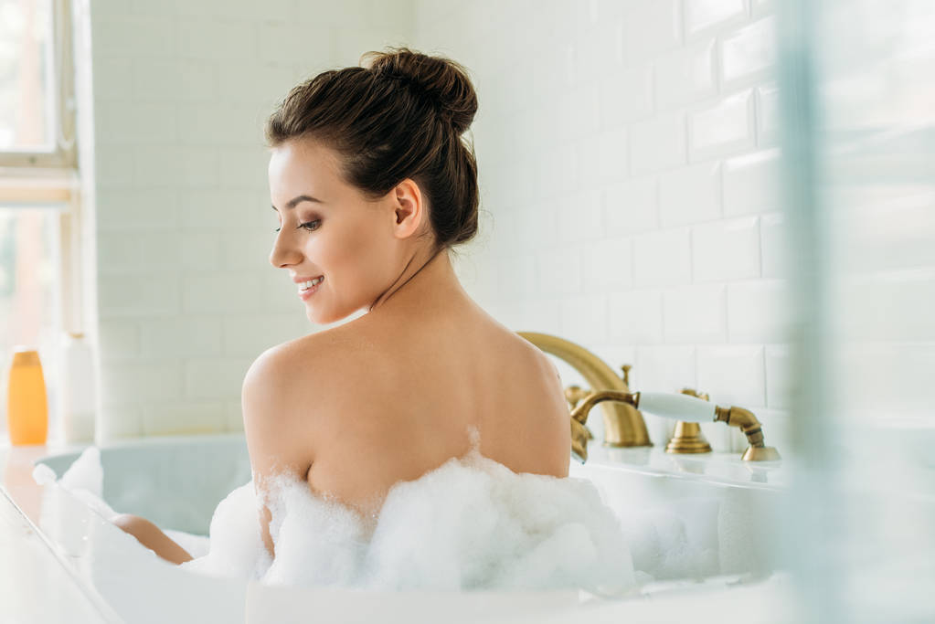 Achteraanzicht van mooie lachende naakt meisje zitten in badkuip met schuim - Foto, afbeelding