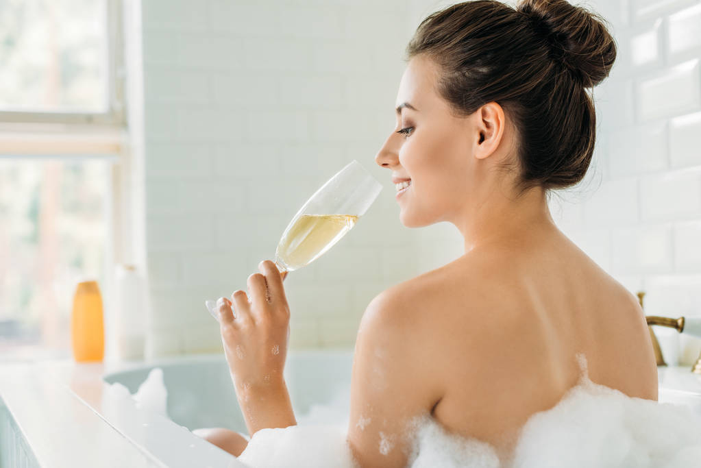 Achteraanzicht van mooi lachende meisje het drinken van wijn in de badkuip met schuim  - Foto, afbeelding