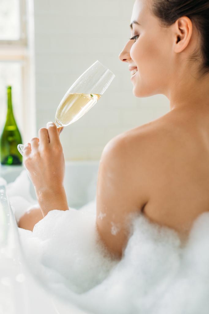 piękna młoda kobieta uśmiechający się picia szampana w wannie z pianki  - Zdjęcie, obraz