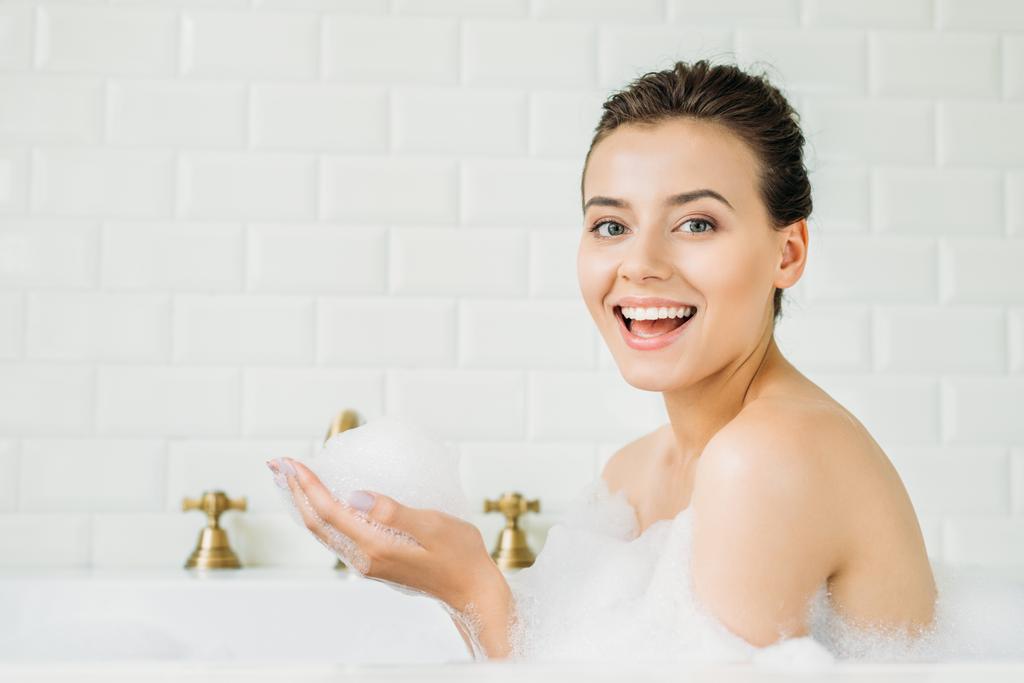 beautiful young woman sitting in bathtub with foam and smiling at camera  - Photo, Image