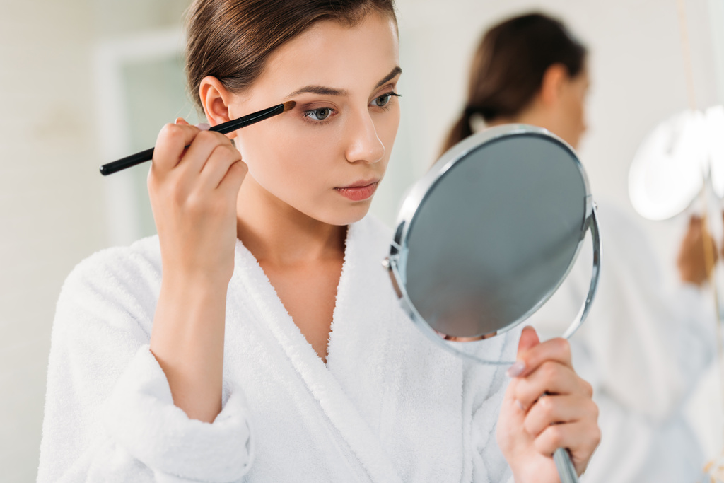 όμορφη νεαρή γυναίκα κρατώντας καθρέφτη και εφαρμόζοντας σκιά ματιών  - Φωτογραφία, εικόνα