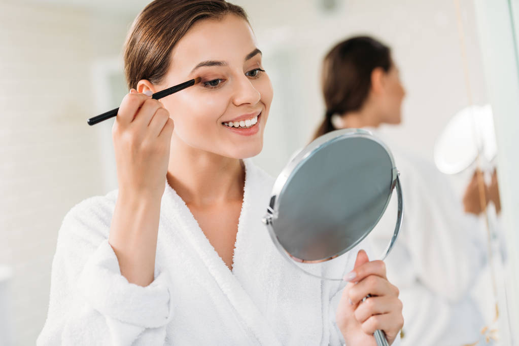 beautiful smiling brunette girl applying makeup in bathroom - Photo, Image