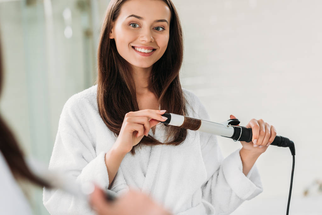 mosolygó fiatal nő a hajsütővas segítségével a fürdőszobában fürdőköpeny  - Fotó, kép