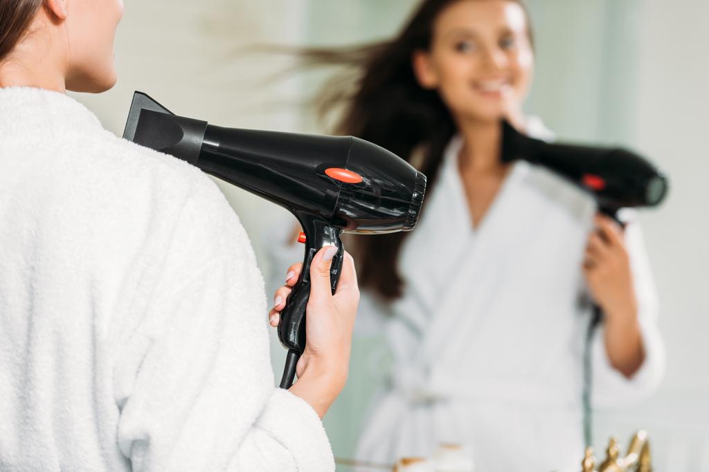 selective focus of smiling girl in bathrobe using hair dryer at mirror in bathroom - Photo, Image
