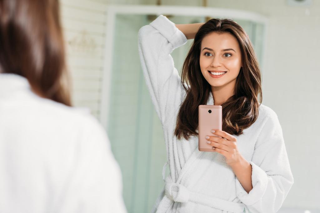 schöne lächelnde junge Frau im Bademantel macht Selfie mit Smartphone im Badezimmer  - Foto, Bild