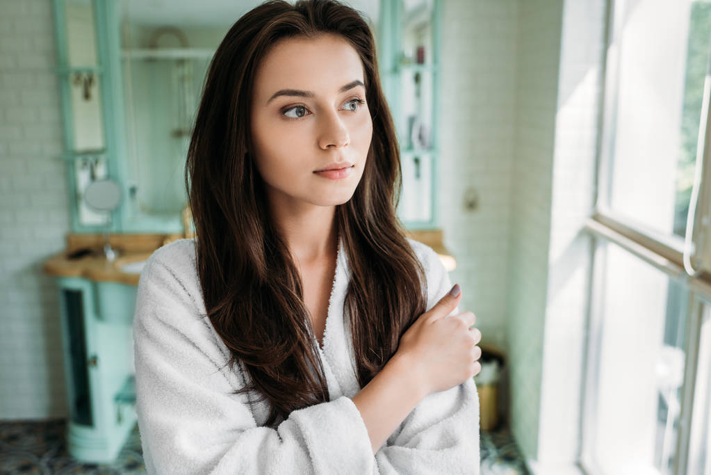 pensive brunette girl in bathrobe looking at window in bathroom - Photo, Image