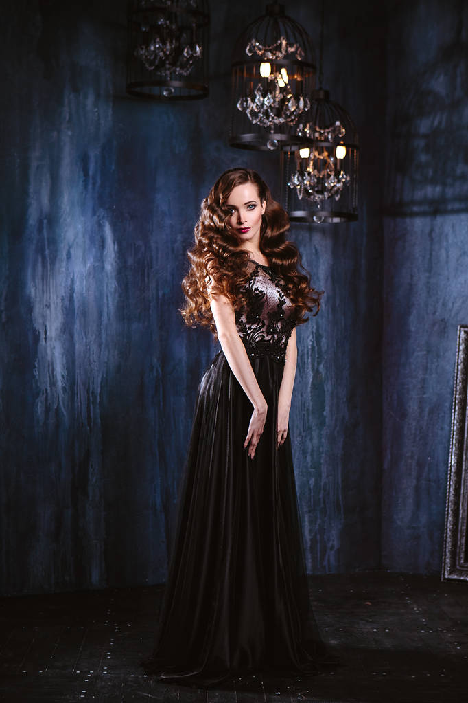 Mladá žena s dlouhé kudrnaté vlasy a make-up večerní dlouhé luxusní šaty, pózuje v temné místnosti interiéru. Módní krása portrét - Fotografie, Obrázek
