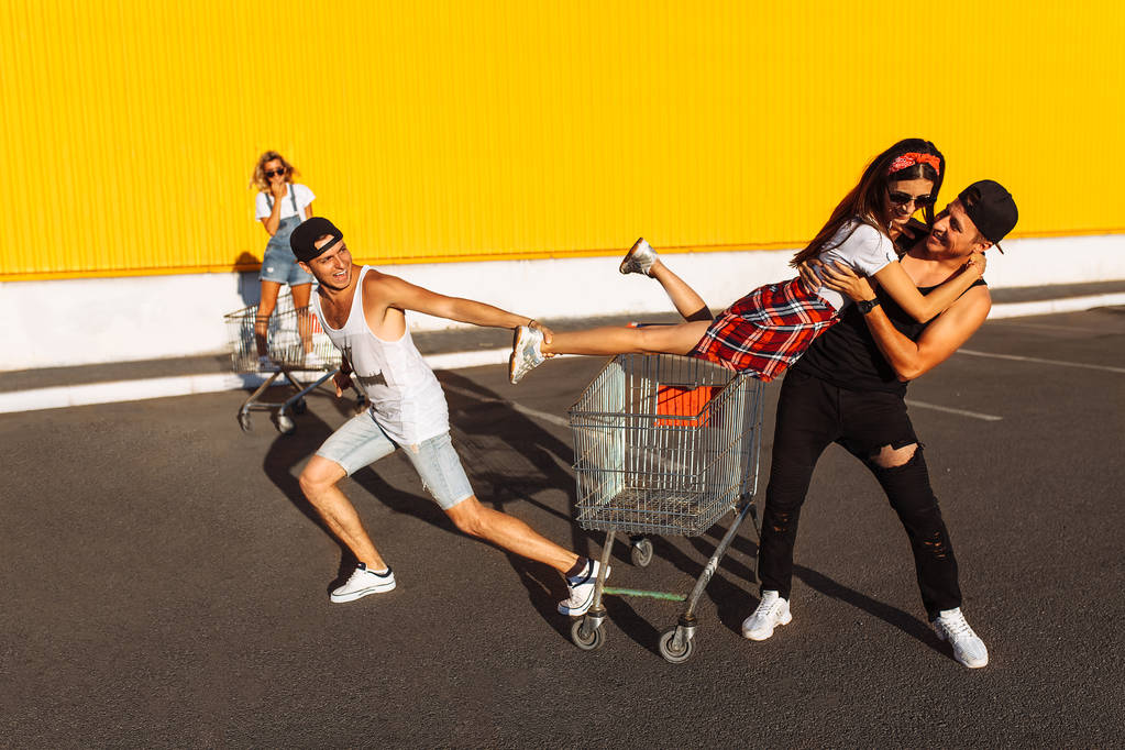 Les amis montent sur des chariots, près du supermarché, un grand passe-temps, un groupe de jeunes monte sur des chariots, les couples amoureux s'amusent par temps ensoleillé
 - Photo, image