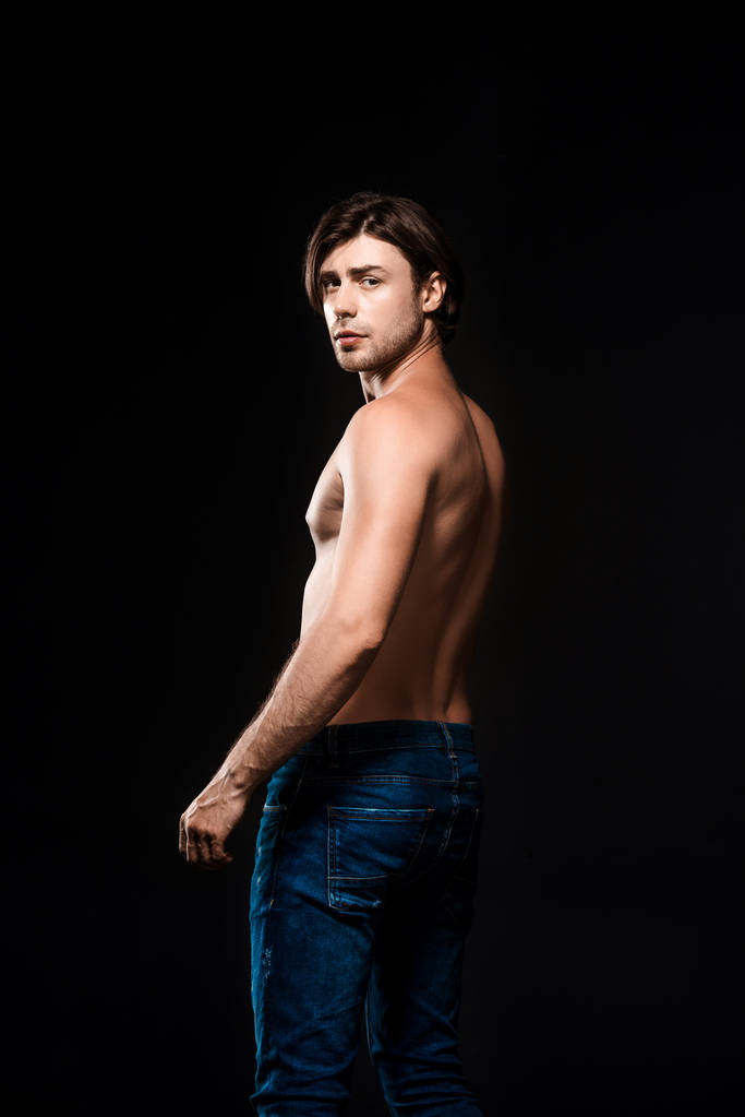 back view of shirtless man in jeans looking at camera isolated on black - Photo, Image