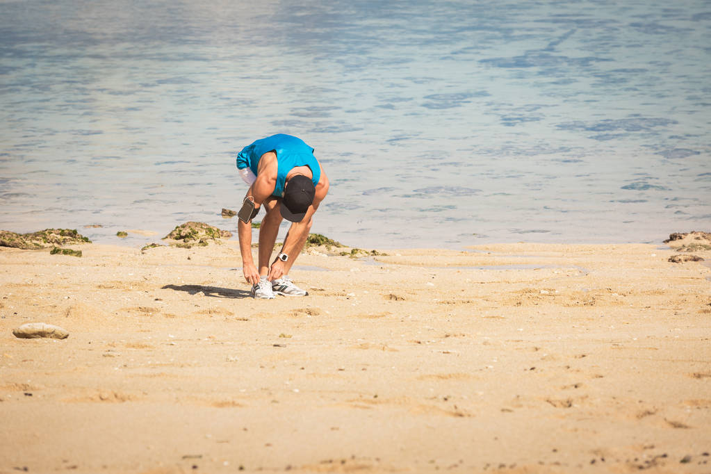 deportista con brazalete de teléfono inteligente atando cordones en la playa cerca del mar
 - Foto, Imagen