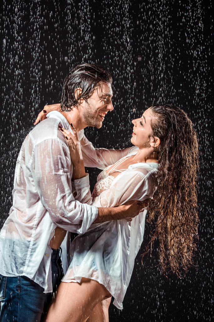 happy wet couple in white shirts standing under rain isolated on black - Photo, Image