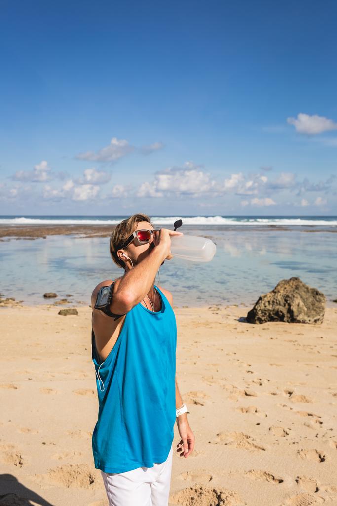 спортсмен пьет воду из спортивной бутылки после тренировки на пляже
 - Фото, изображение