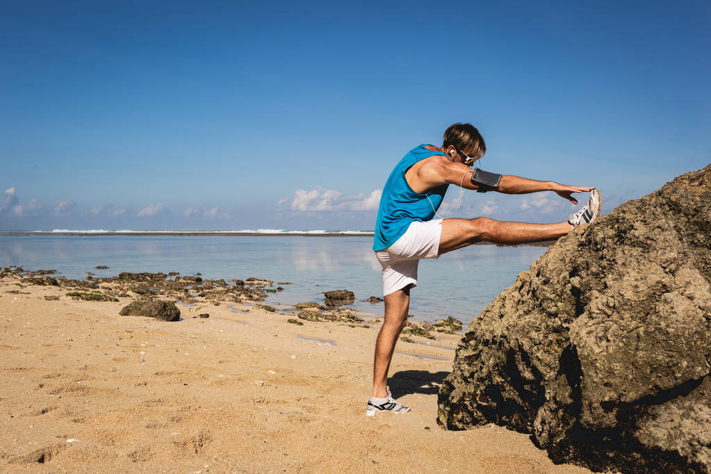 lekkoatletycznego mężczyzna stretching nóg w pobliżu skały na plaży - Zdjęcie, obraz