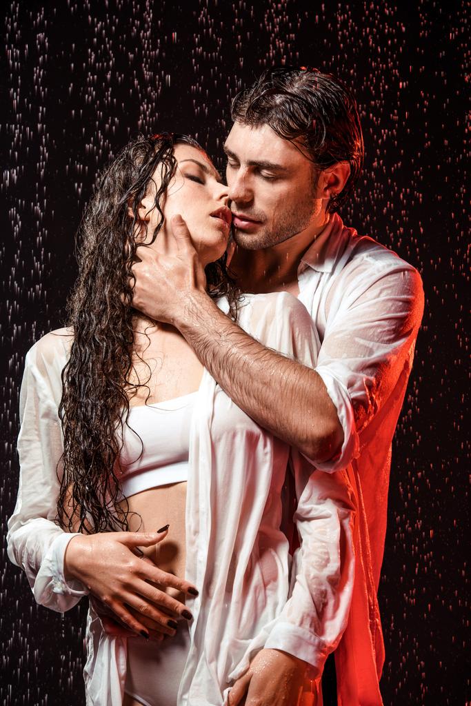 Porträt eines sexy Paares in weißen Hemden, das unter Regen vor schwarzem Hintergrund steht - Foto, Bild