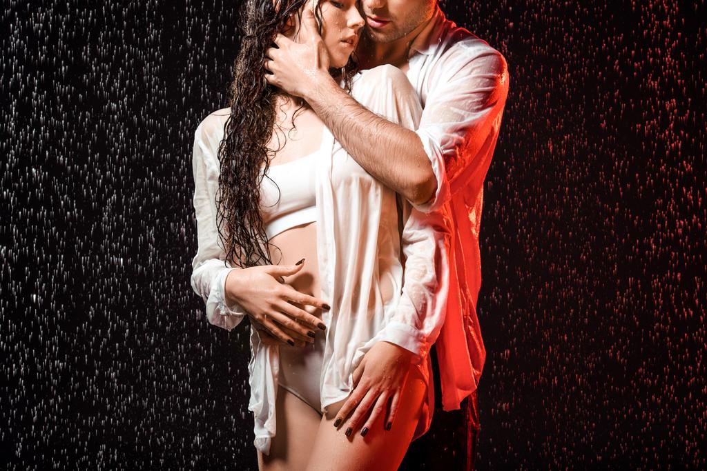 osittainen näkymä seksikäs pari valkoisissa paidoissa seisoo sateen alla mustalla taustalla
 - Valokuva, kuva