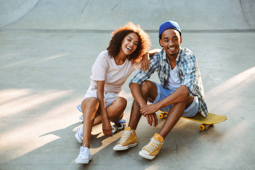 Retrato de um jovem casal africano sorridente com skates sentados juntos no parque de skate
 - Foto, Imagem