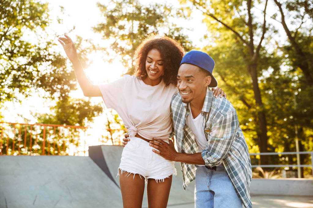 Retrato de una alegre pareja africana joven con monopatines divirtiéndose juntos en el parque de skate, enseñando a montar
 - Foto, imagen