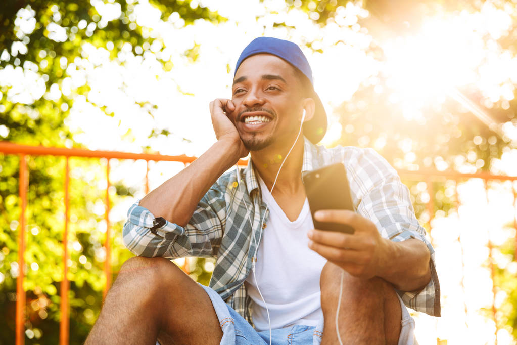 Joven africano alegre con auriculares escuchando música con teléfono móvil en una rampa en el parque de skate
 - Foto, Imagen
