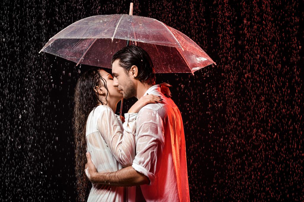 side view of romantic couple in white shirts with umbrella standing under rain on black backdrop - Photo, Image