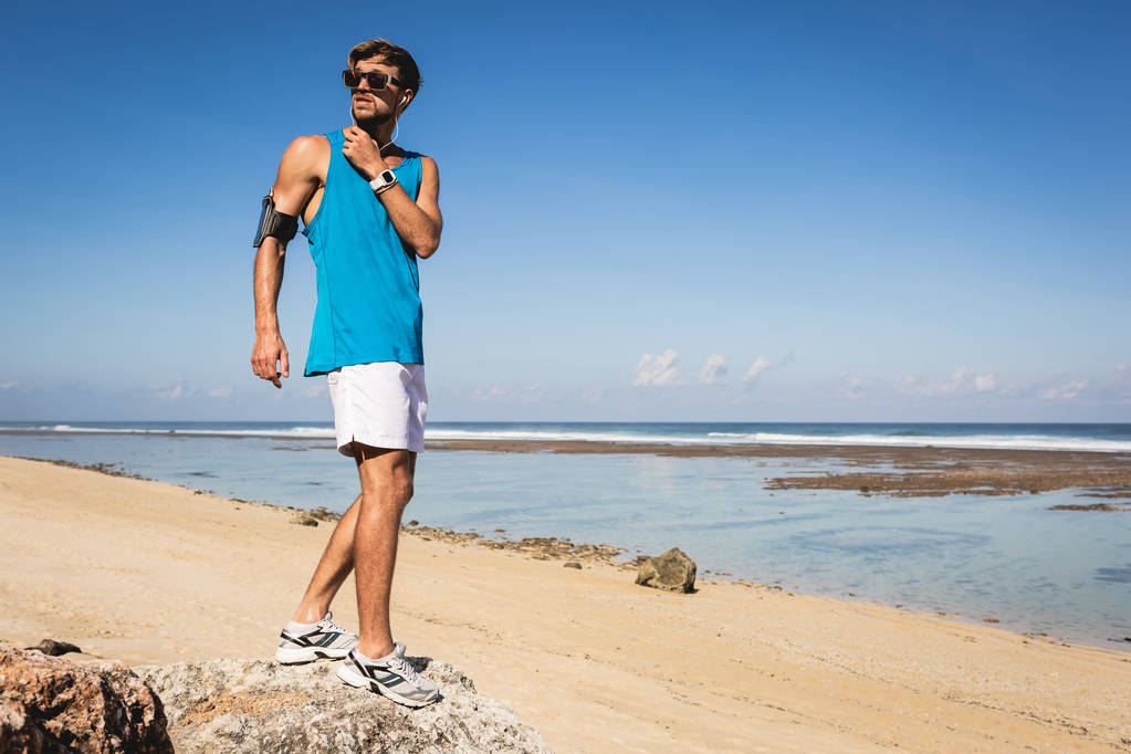 deportista escuchando música con smartphone en brazalete mientras está de pie en la playa, Bali, Indonesia
 - Foto, imagen