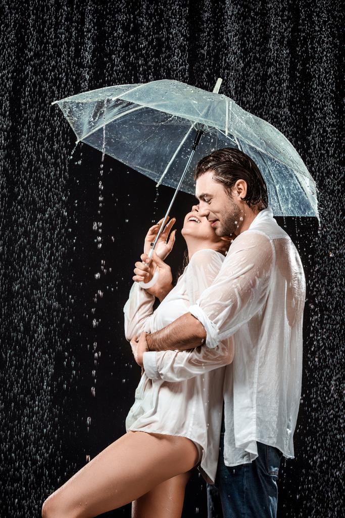 Widok z boku z romantyczna para w białe koszule, stojąc pod parasol w deszczu na czarnym tle - Zdjęcie, obraz
